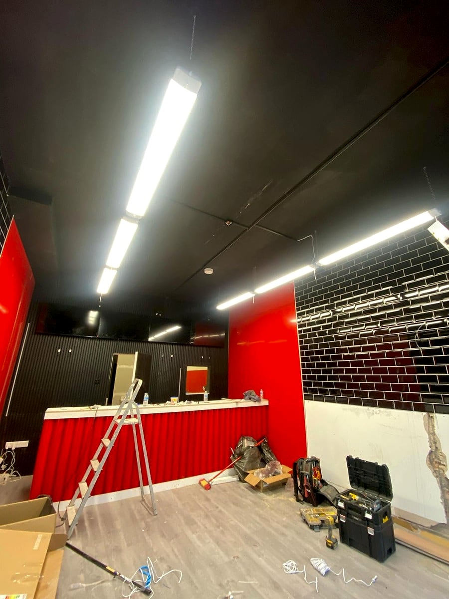 Suspended linear LED lights on over the black ceiling at Cheat Daze, with the red counter and black tiled wall below