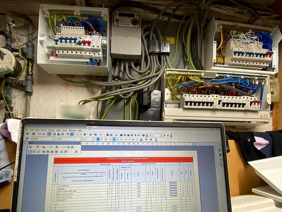 Consumer units opened up for an EICR, with the schedule of test results being filled in on a laptop